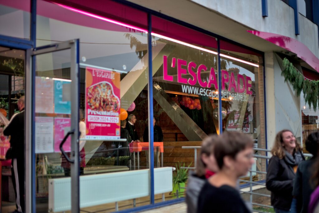 Façade du théâtre de l'Escapade depuis l'extérieur. En premier plan, floues, des personnes discutent.