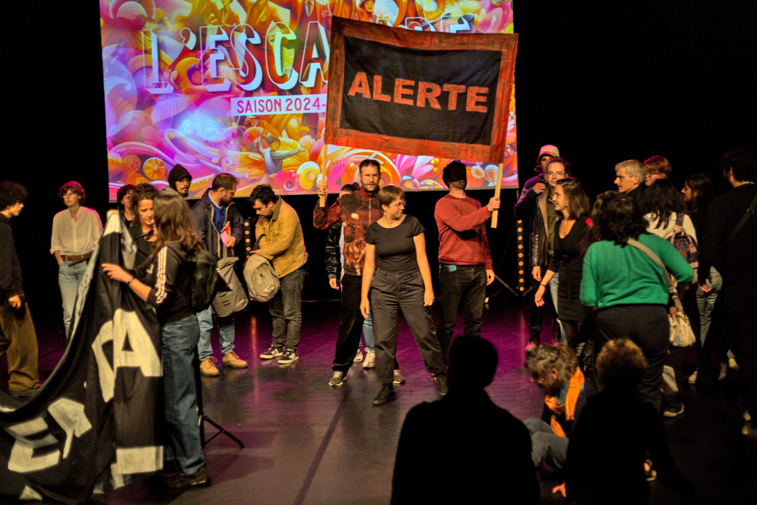 Action sur les planches de la scène principale du théâtre. Plusieurs militant-es montent sur scène. Certain-es tiennent une banderole "ALERTE".