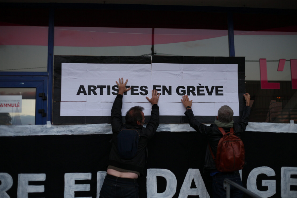 2 militants tentent d'accrocher une grande pancarte "ARTISTES EN GREVE" sur les vitres de l'entrée du théâtre.